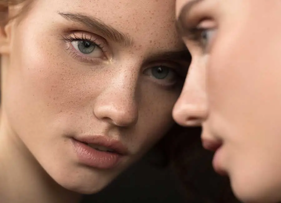 a close up image of a freckled woman with smooth skin leaning against a mirror while staring at the camera
