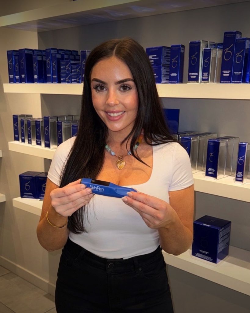 Woman with long dark hair in a white top smiling while holding a blue SkinPen box in a skincare shop.